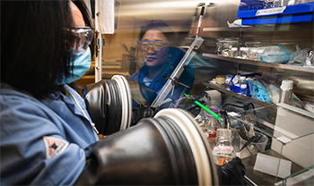 battery scientist in lab gear reaches into closed machine with attached gloves as another person is seen in reflection