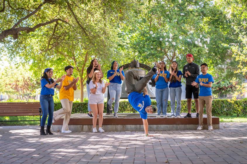 Students hanging out and dancing on campus. Greenery surrounds them with tower bell right behind them.